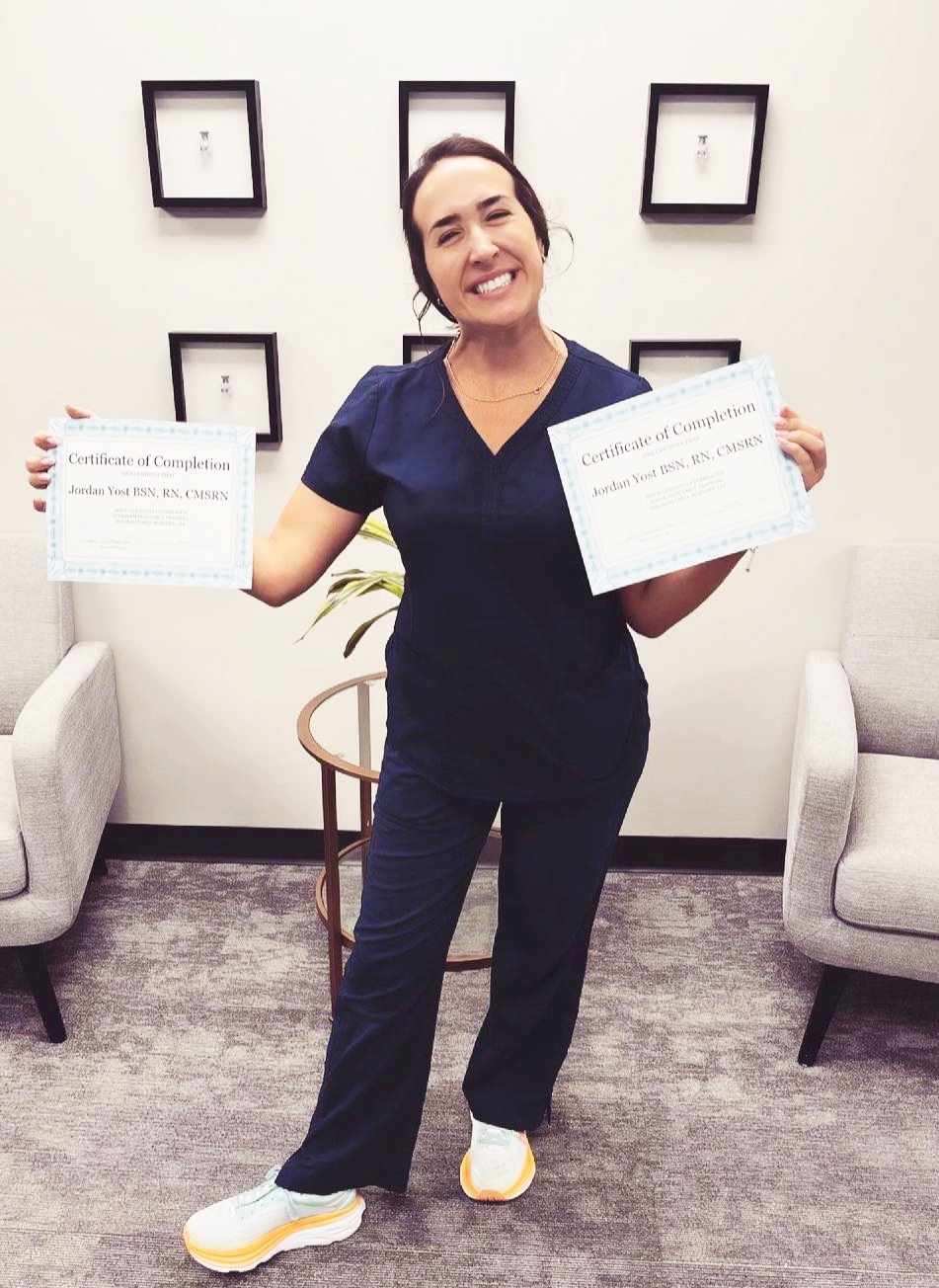 Jordan Yost wearing navy blue scrubs, holding up two professional certificates, smiling.